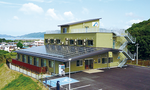 黄色い外壁の介護施設の建物外観。駐車場と周辺の山々が見える。